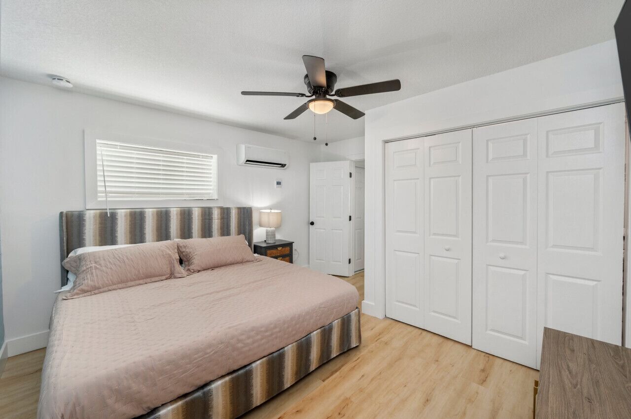 A bedroom with a bed, a ceiling fan, a window with blinds, an air conditioning unit, and a set of white doors.