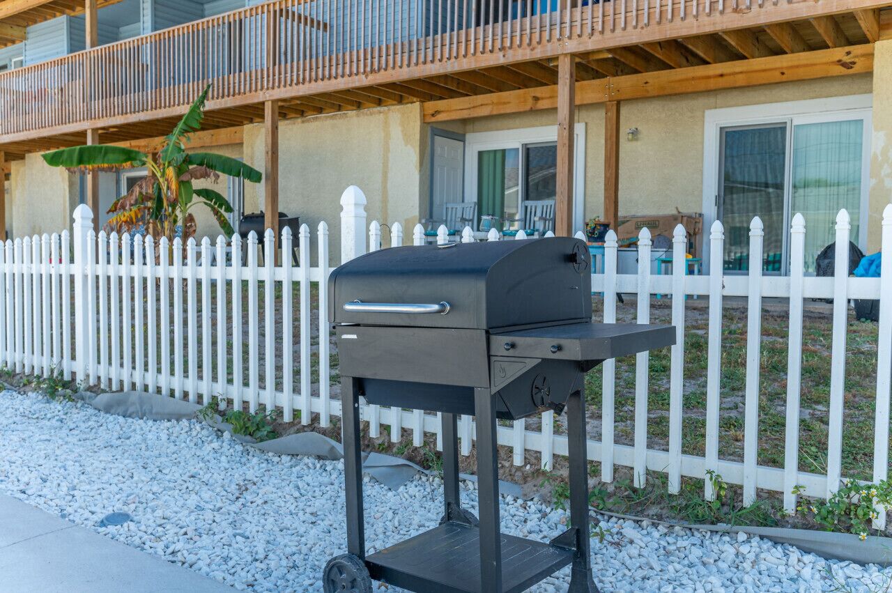 A portable grill in front of a white picket fence with a balcony above.