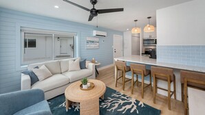Un salon moderne avec un canapé, une table basse ronde et un ventilateur de plafond. Il y a également un coin cuisine avec un comptoir-bar et des tabourets.