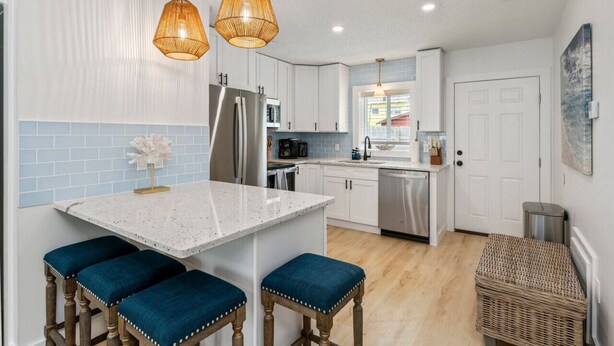 Cocina moderna con armarios blancos, una isla central con taburetes azules, electrodomésticos de acero inoxidable y cestas de mimbre.