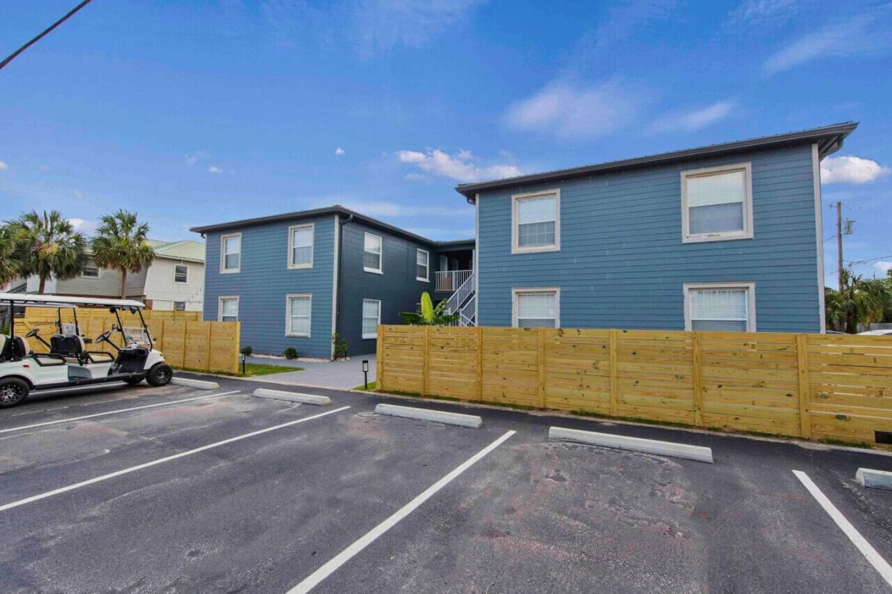 Un parking avec un véhicule de golf, une clôture en bois et deux bâtiments bleus dotés de fenêtres et d’un balcon.