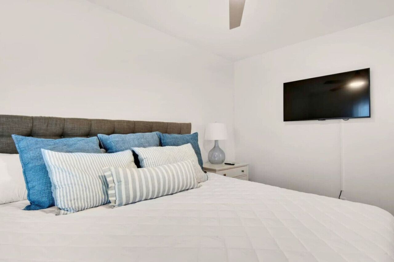 Un dormitorio con cama, almohadas, mesita de noche, lámpara y televisor montado en la pared.