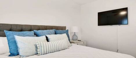Un dormitorio con cama, almohadas, mesita de noche, lámpara y televisor montado en la pared.