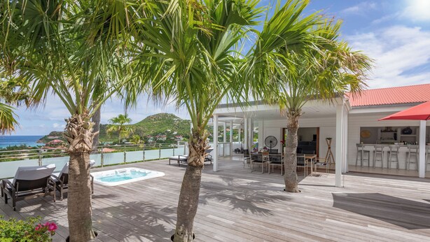 Une terrasse en bois avec une piscine, un espace salon extérieur et une zone couverte avec des tables et des chaises.