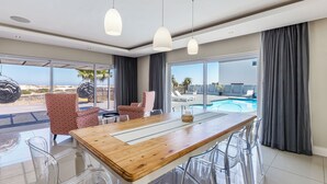 Ein moderner Essbereich mit Holztisch, klaren Stühlen und Pendelleuchten. Große Fenster bieten Blick auf einen Pool und Sitzgelegenheiten im Freien.
