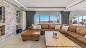 Ein modernes Wohnzimmer mit Kamin aus Ziegelsteinen, Ledersofas, einem Holztisch und großen Fenstern mit Blick auf den Strand.