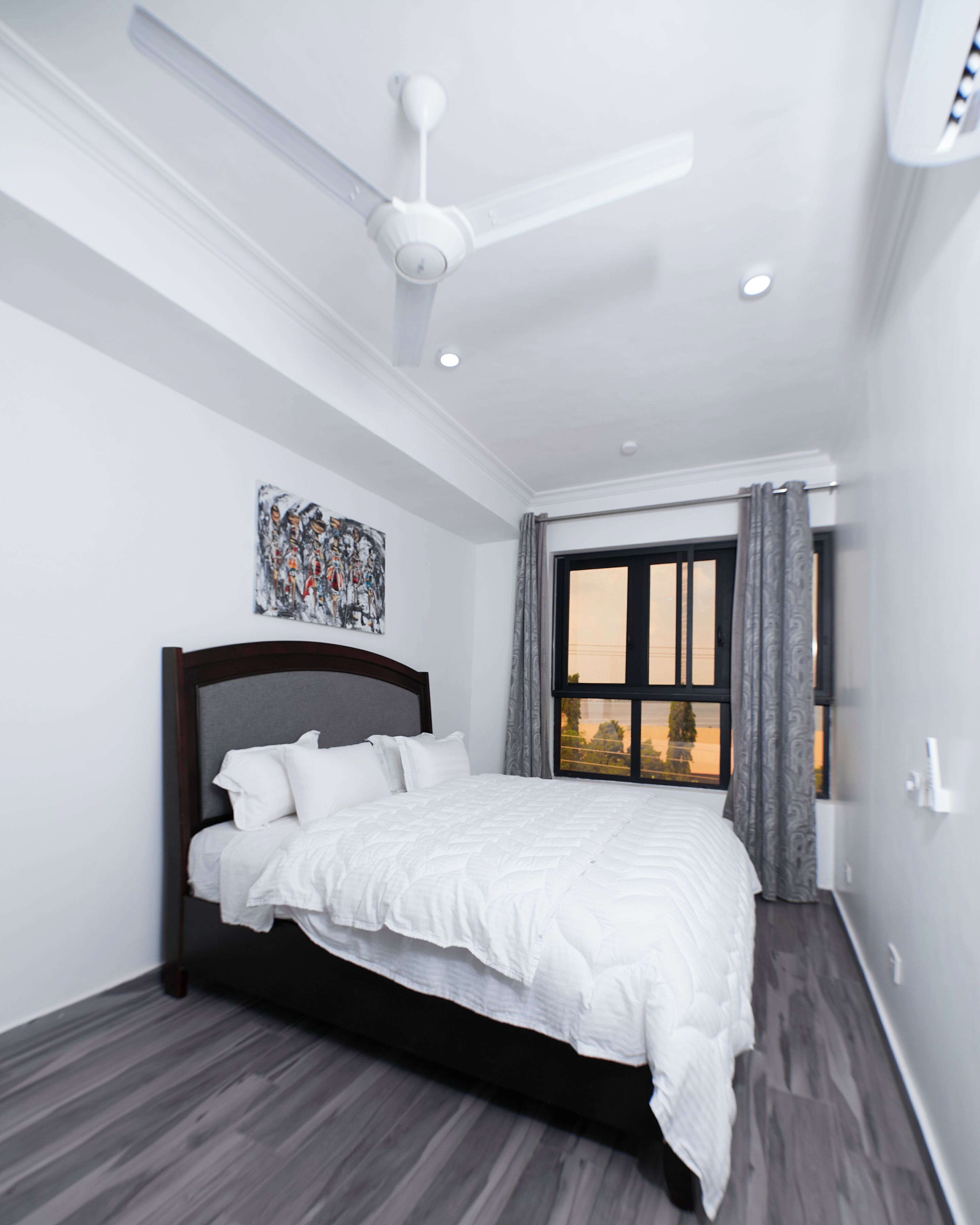 Un dormitorio moderno con una cama, un ventilador de techo, una ventana con cortinas y un cuadro en la pared.