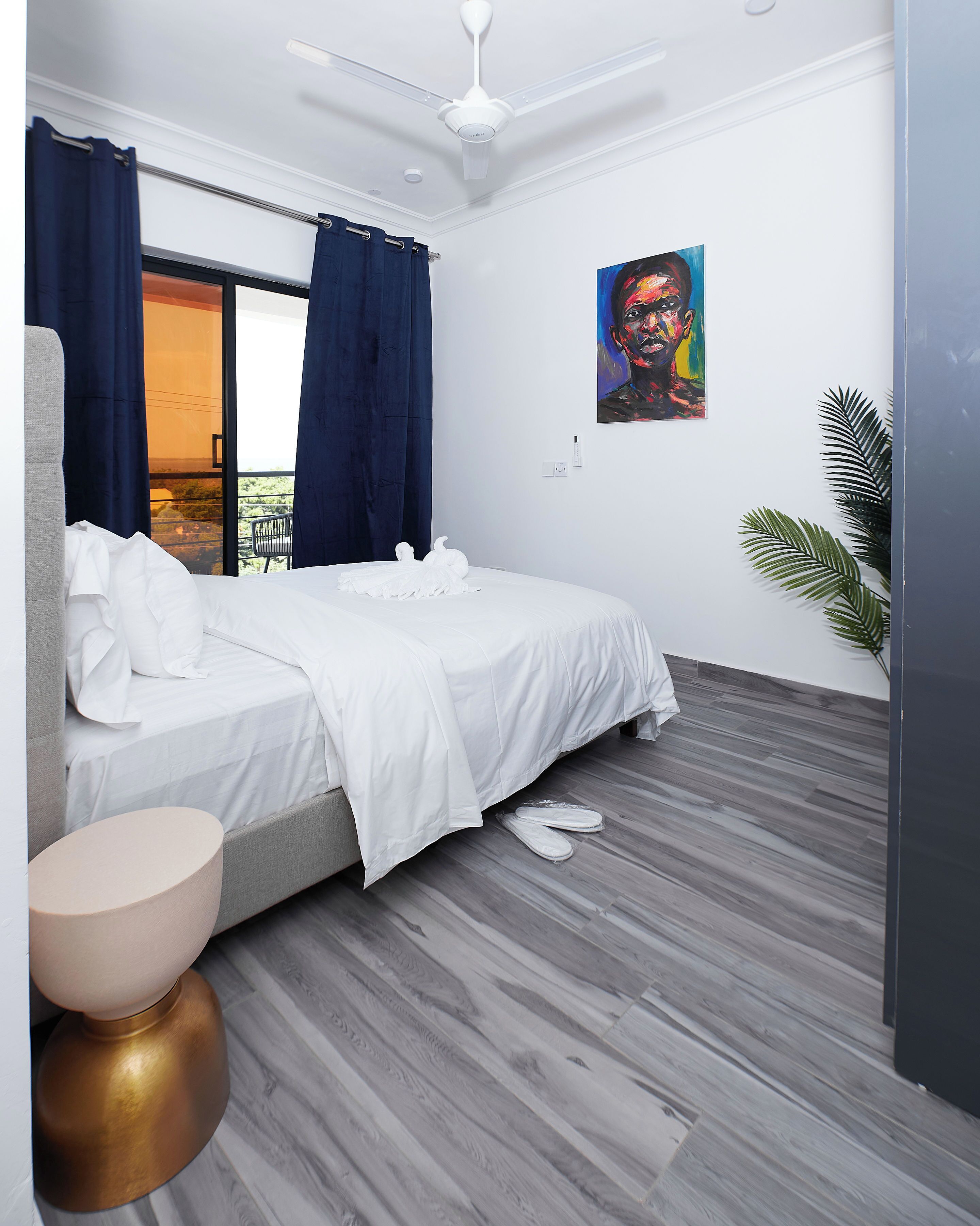 A modern bedroom with a bed, a ceiling fan, a window with curtains, a painting on the wall, and a potted plant.