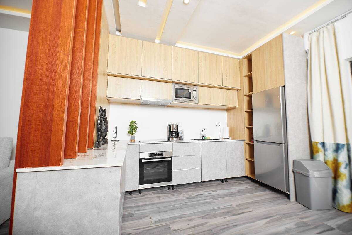 A modern kitchen with wooden cabinets, a microwave, a built-in oven, a refrigerator, and a trash bin.