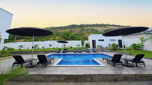 A swimming pool with lounge chairs and umbrellas, surrounded by a landscaped garden and a hillside in the background. - River Hills Hotel & Suites (Mile 11)