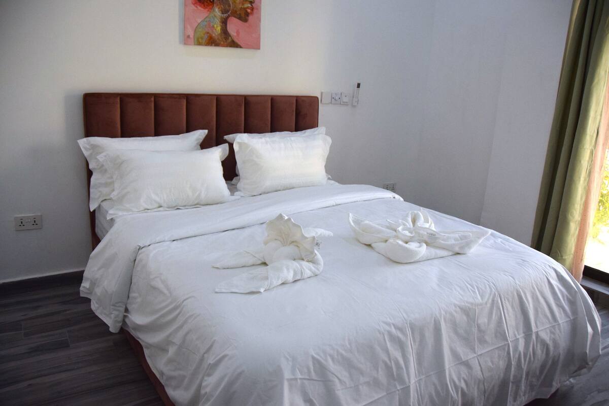 A neatly made bed with white linens and two pillows, a brown headboard, and a painting on the wall.