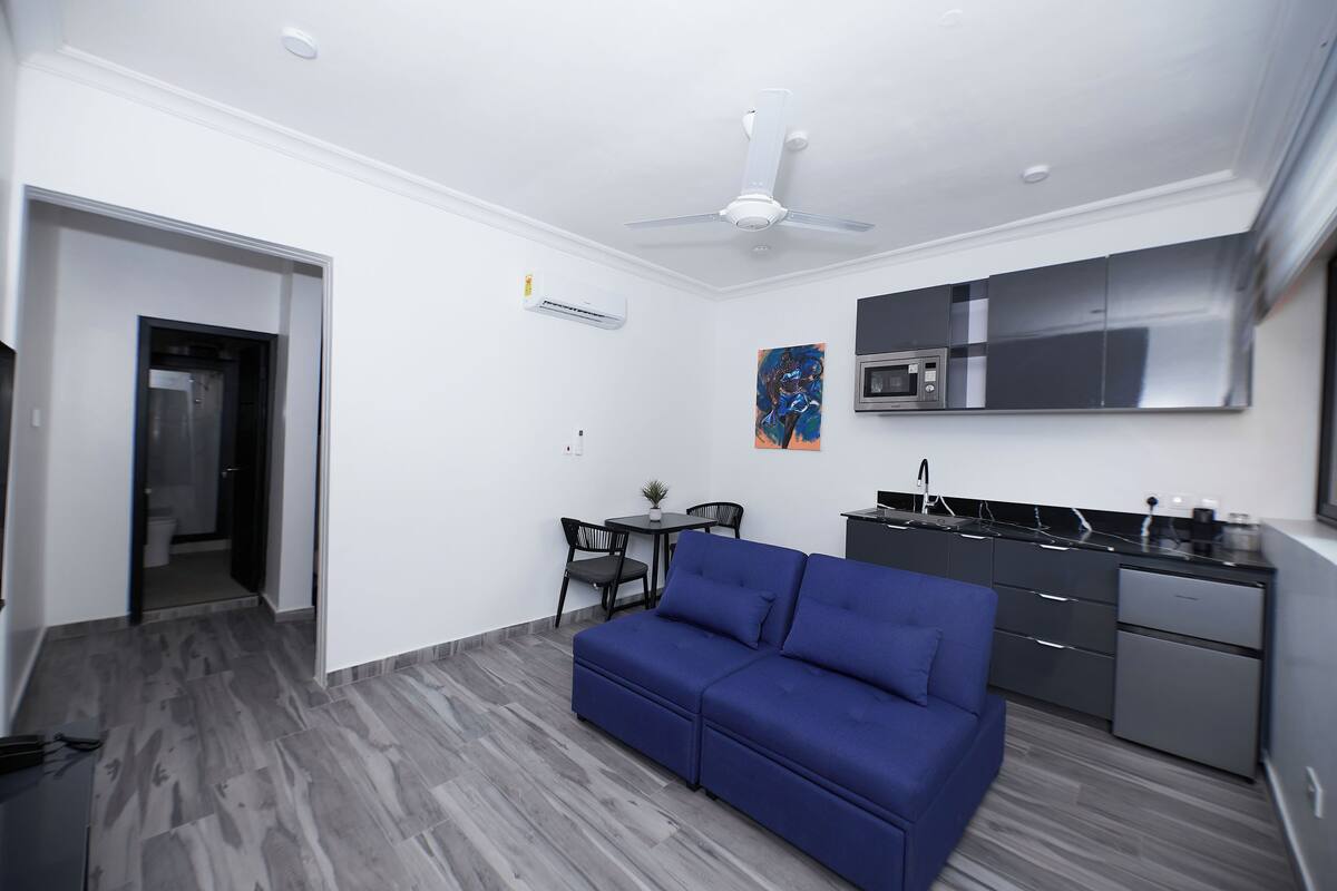 A modern living room with a blue sofa, a dining area with a small table and chairs, a kitchen with a microwave and cabinets, and a ceiling fan.