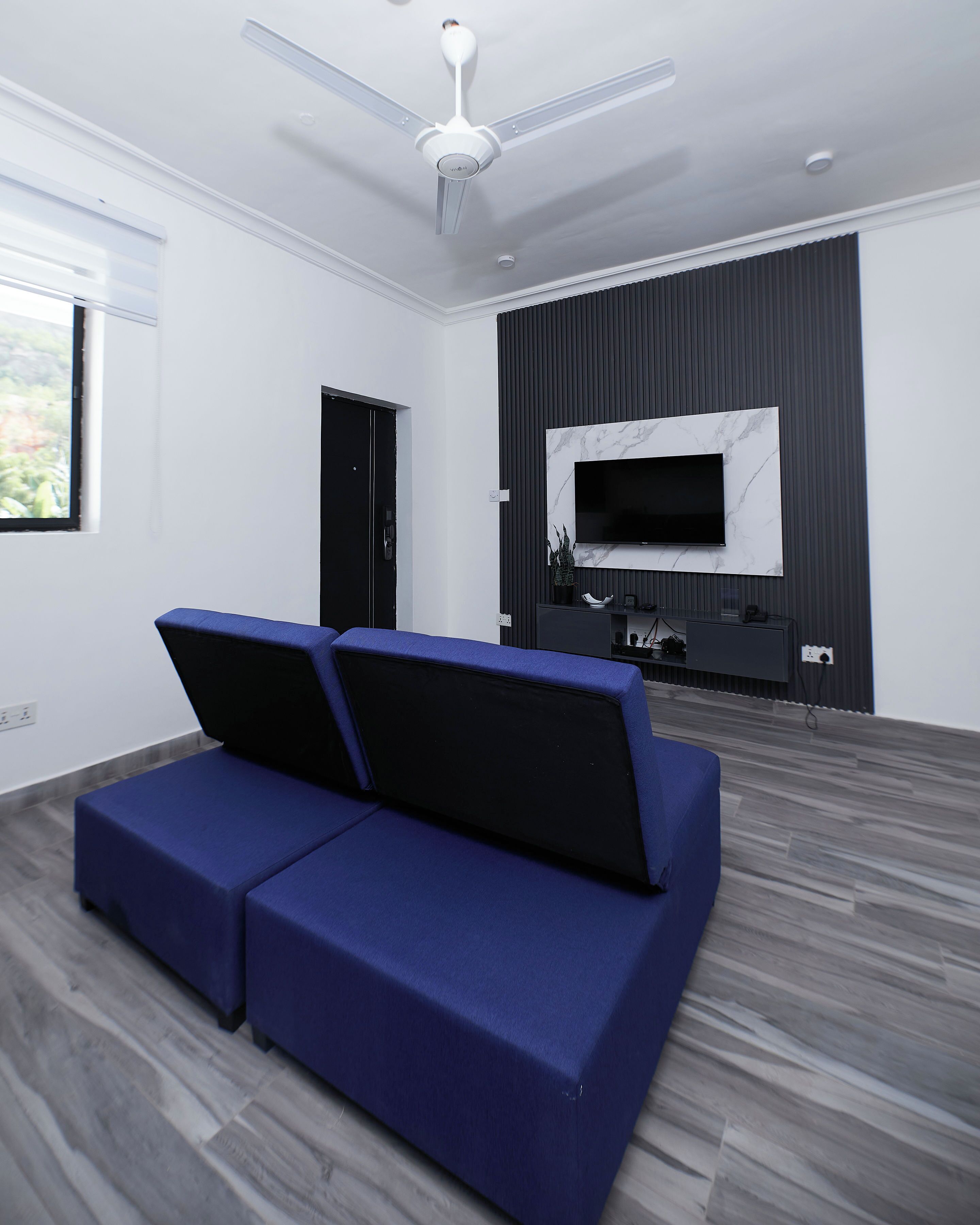 A modern living room with a blue sofa, a flat-screen TV mounted on a dark wall, and a ceiling fan.
