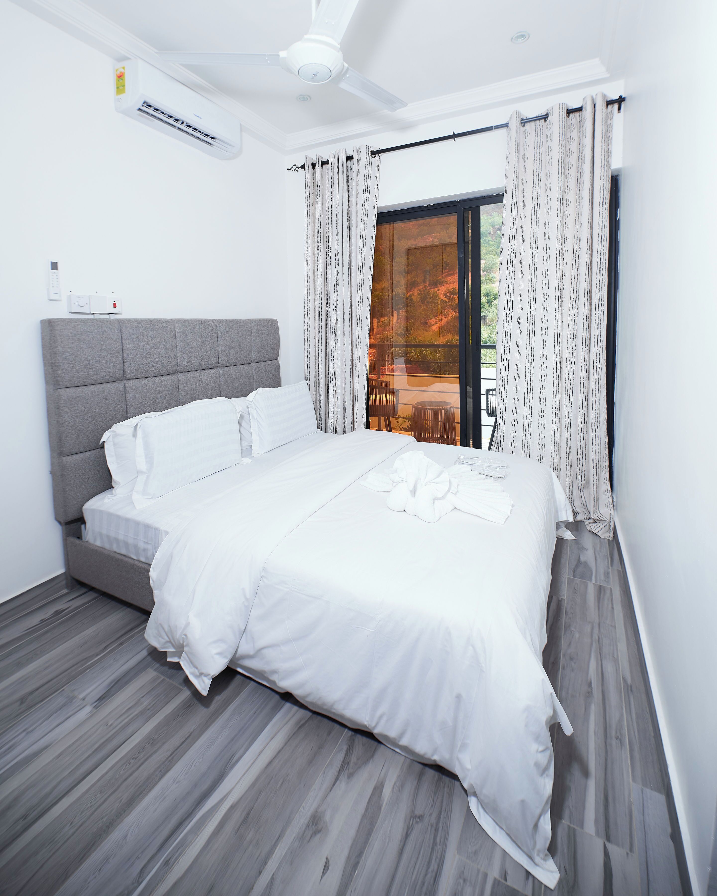 A modern bedroom with a grey upholstered headboard, a white bedspread, and a large window with patterned curtains.