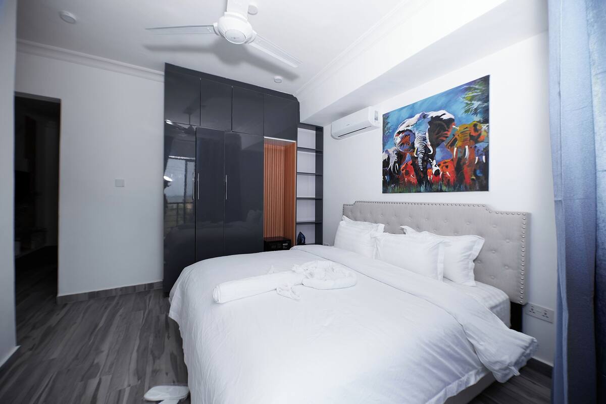 A modern hotel room with a large bed, a ceiling fan, a wardrobe, and a painting on the wall.