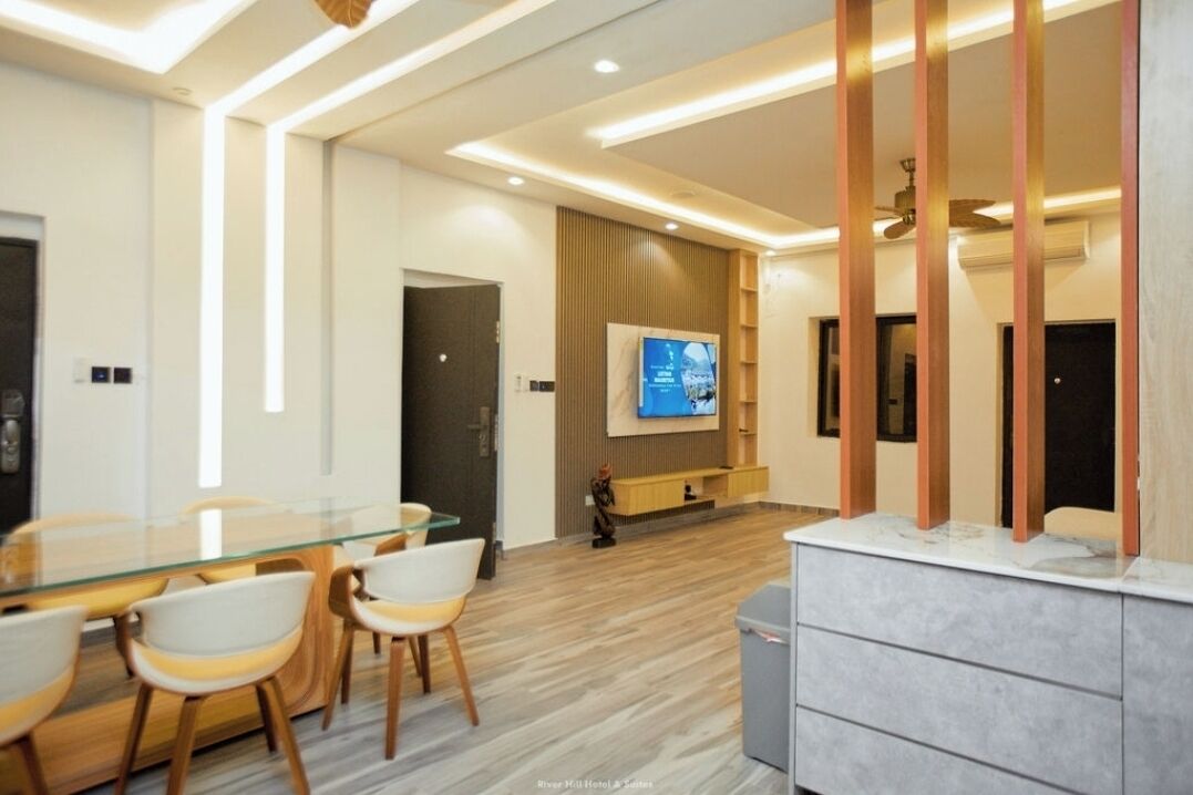 A modern hotel room with a glass dining table, white chairs, a flat-screen TV mounted on the wall, and a wooden cabinet.