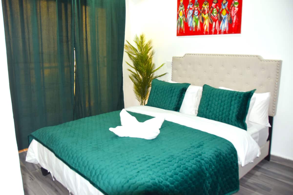 A neatly made bed with a green bedspread and white linens, a grey headboard, and a decorative pillow.