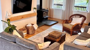 A cozy living room with a flat-screen TV, a fireplace, wicker furniture, and a large window with curtains.