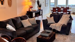 A cozy living room with two dark sofas, a wicker chair, a coffee table, and a rug. There is a dining area with wooden chairs and a table in the background.