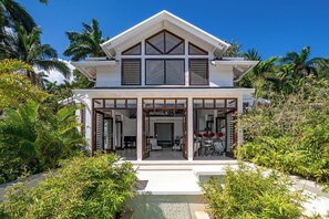 Una casa de dos pisos con un porche cubierto, ventanas grandes y un jardín bien cuidado con plantas tropicales.