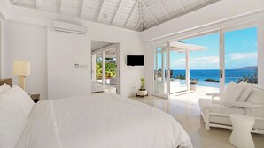 A spacious bedroom with a large bed, a ceiling fan, and a TV. It features a balcony with a view of the ocean.