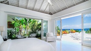 A modern bedroom with a large bed, white furniture, and a view of the ocean through large sliding doors.