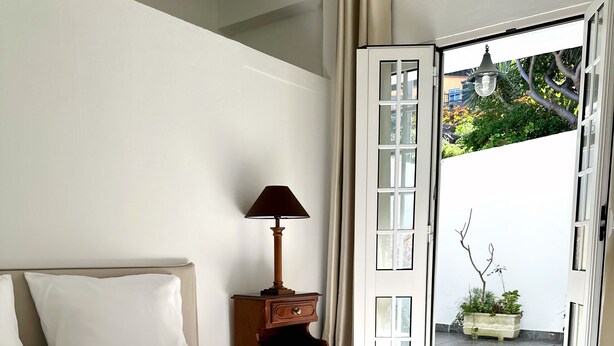 A hotel room with a bed, white pillows, a wooden nightstand with a lamp, and a view of a garden through an open door. - K&K Apartments (Machico)