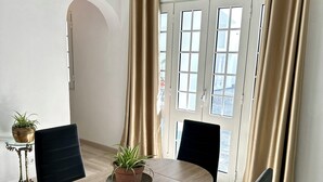 A modern dining area with a round table, four black chairs, a potted plant, and a view through a large window with beige curtains. - K&K Apartments (Machico)