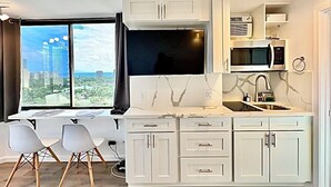A modern kitchen with white cabinetry, a large window with a city view, a dining area with two chairs, and a built-in microwave. - Beautiful OCEAN & CANAL View - Renovated - Waikiki Studio (Honolulu)