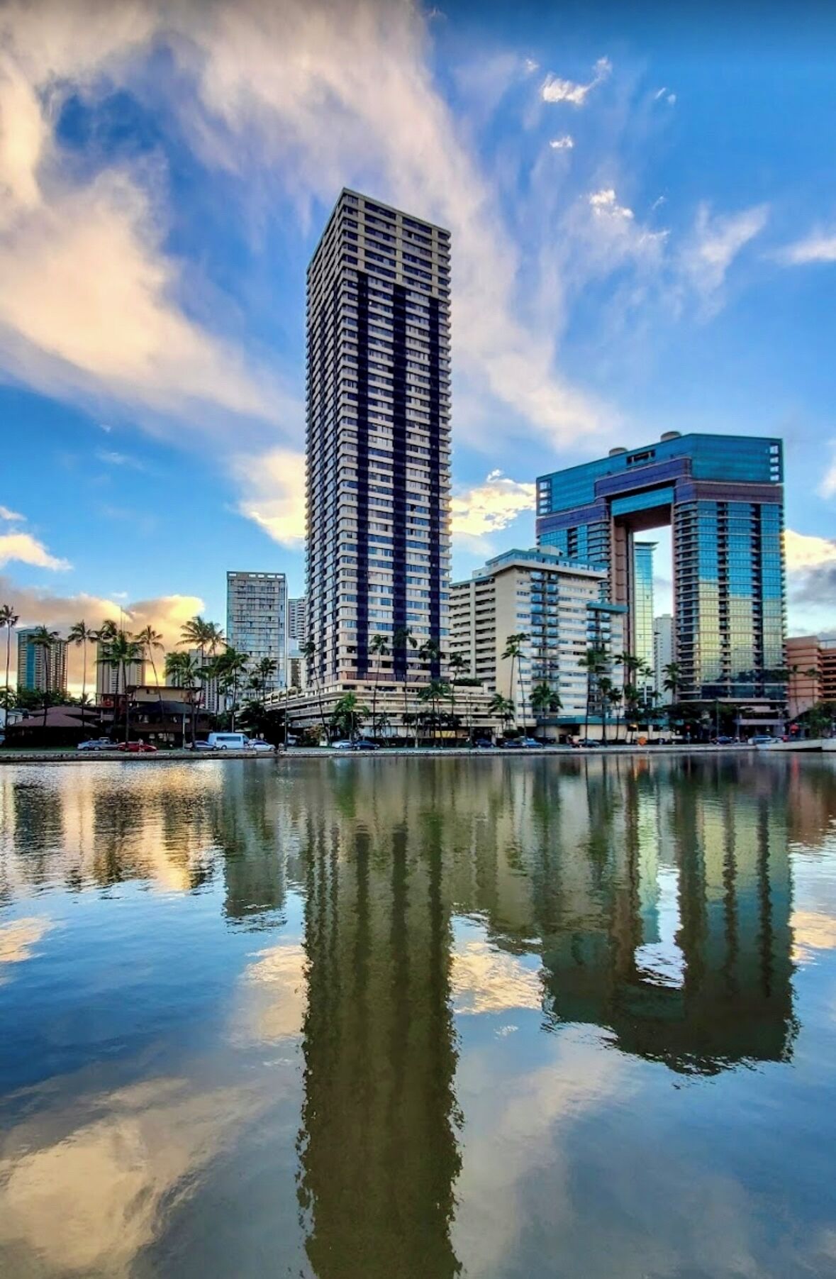 A cityscape with a tall building reflecting in a calm body of water, surrounded by other high-rise buildings.