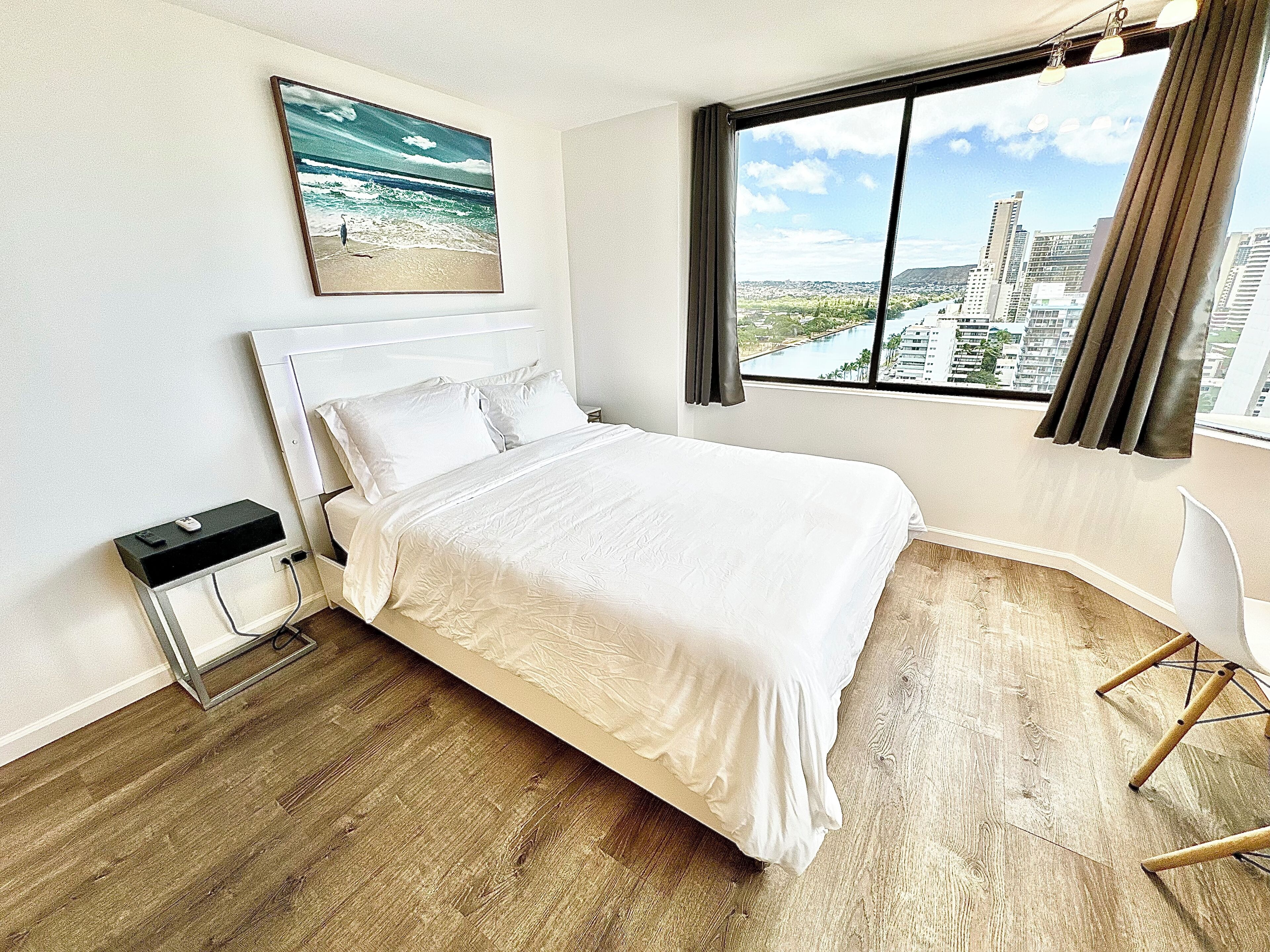 Una habitación de hotel con cama, mesita de noche, silla, ventana con vista a la ciudad y un cuadro en la pared.