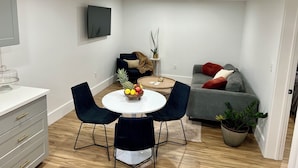 A modern living room with a dining area, a grey sofa, a TV mounted on the wall, and a small table with a fruit bowl.