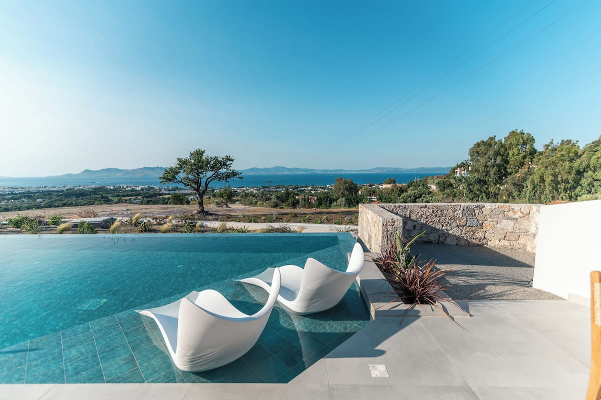 Une magnifique piscine à débordement d’un bleu profond, agrémentée de chaises blanches au design moderne, d’un mur en pierre et offrant une vue sur la mer et les montagnes.