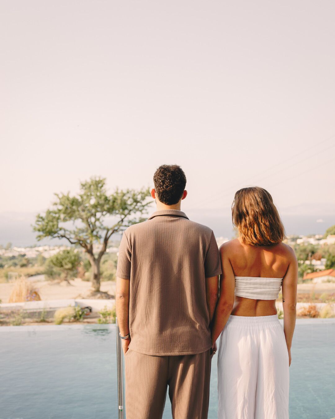 Un couple se tient près d’une piscine, avec vue sur un arbre et des bâtiments situés au loin.