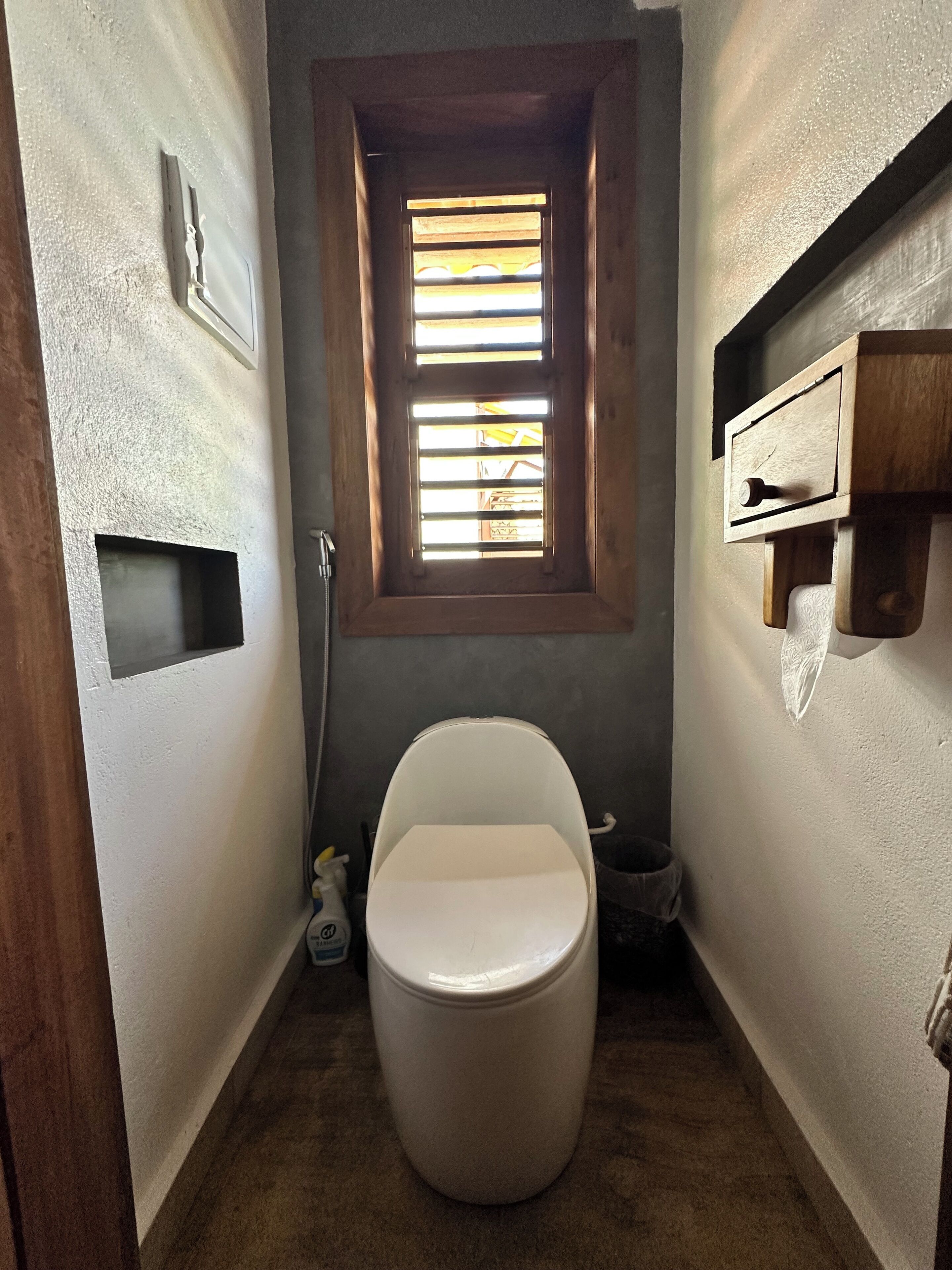 Ein kleines Badezimmer mit Toilette, einem Fenster mit Jalousien, einer Dusche und einem Holzregal.