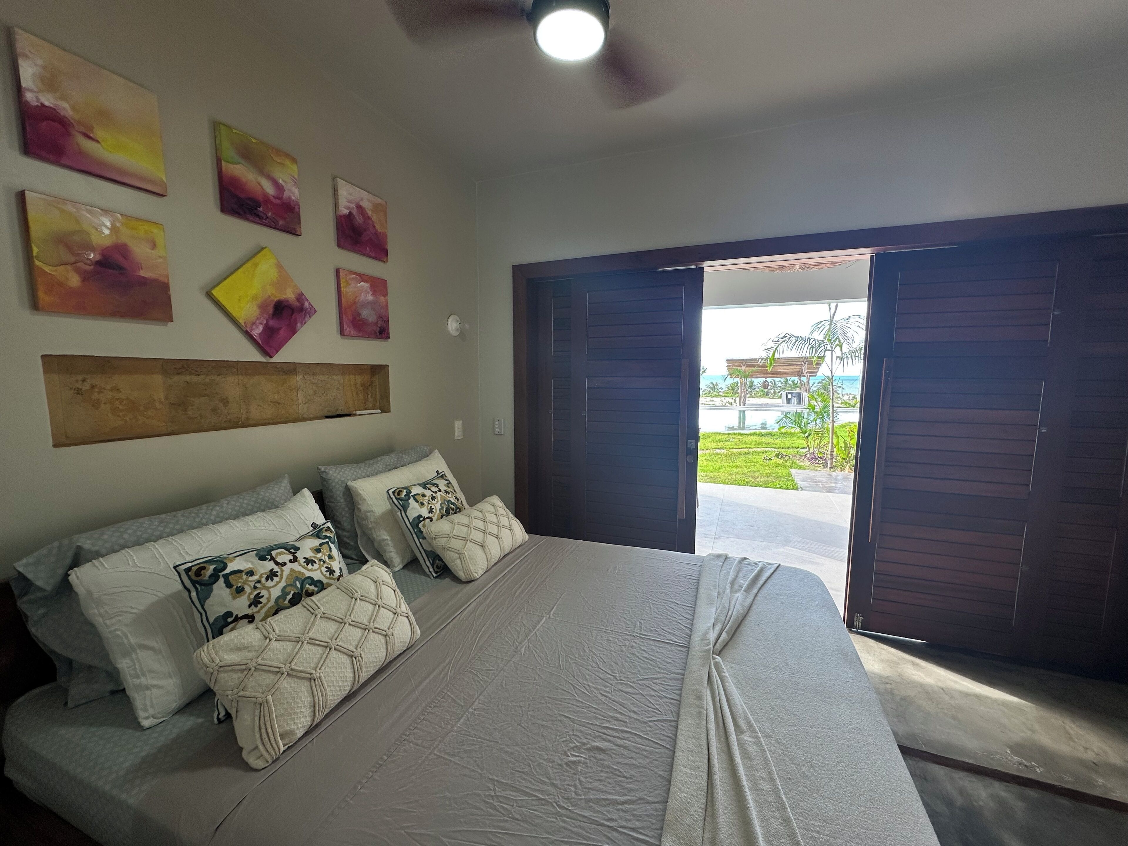 Quarto com cama, almofadas e vista para uma piscina e vegetação.