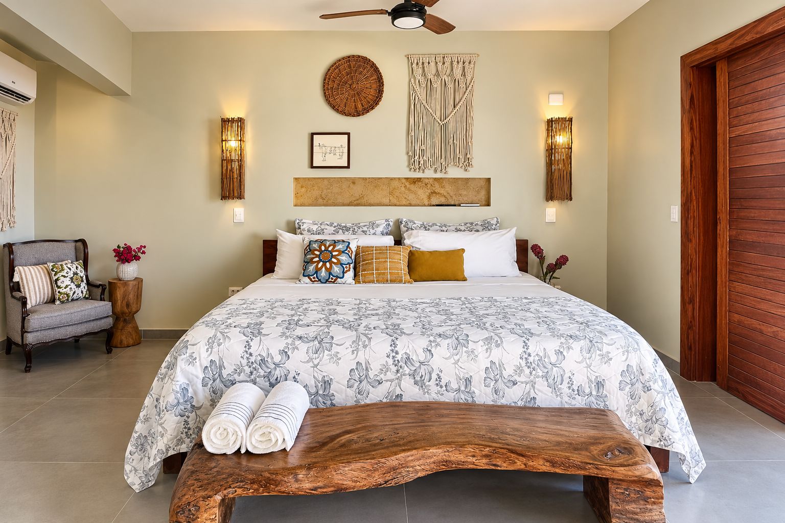 Un dormitorio con una cama grande, un banco de madera, una silla y una mesita de noche con un florero.