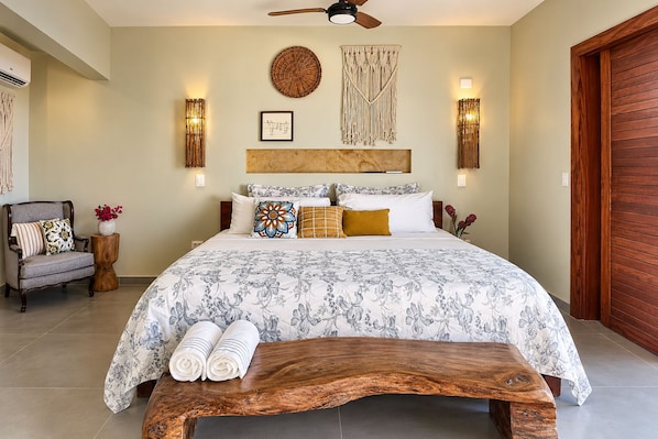 A bedroom with a large bed, a wooden bench, a chair, and a bedside table with a vase of flowers. - Refugio Dunas prea (Cruz)