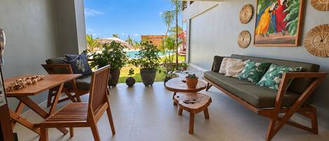 A modern living room with a sofa, chairs, a coffee table, and a dining table. There is a large window providing a view of a pool and greenery.