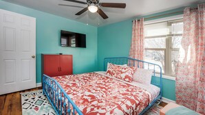 Ein Schlafzimmer mit Bett, Deckenventilator, Fenster mit Vorhängen, einem roten Kleiderschrank und einem an der Wand montierten Fernseher.