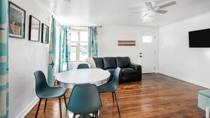 Ein modernes Wohnzimmer mit einem runden Esstisch, blaugrünen Stühlen, einem schwarzen Ledersofa und einem Deckenventilator.