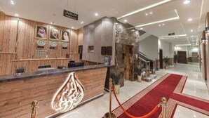 Un hall d’hôtel avec un comptoir d’accueil en bois, des portraits encadrés, une moquette rouge et un sol en marbre.