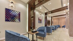 Un hall d’hôtel avec des fauteuils à revêtement bleu, une table en bois sur laquelle se trouve une boîte de mouchoirs, et un tableau abstrait aux couleurs vives accroché au mur.