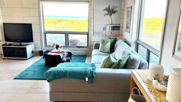 A living room with a sofa, a television, a window with a view of the beach, and a small table with decorative items.