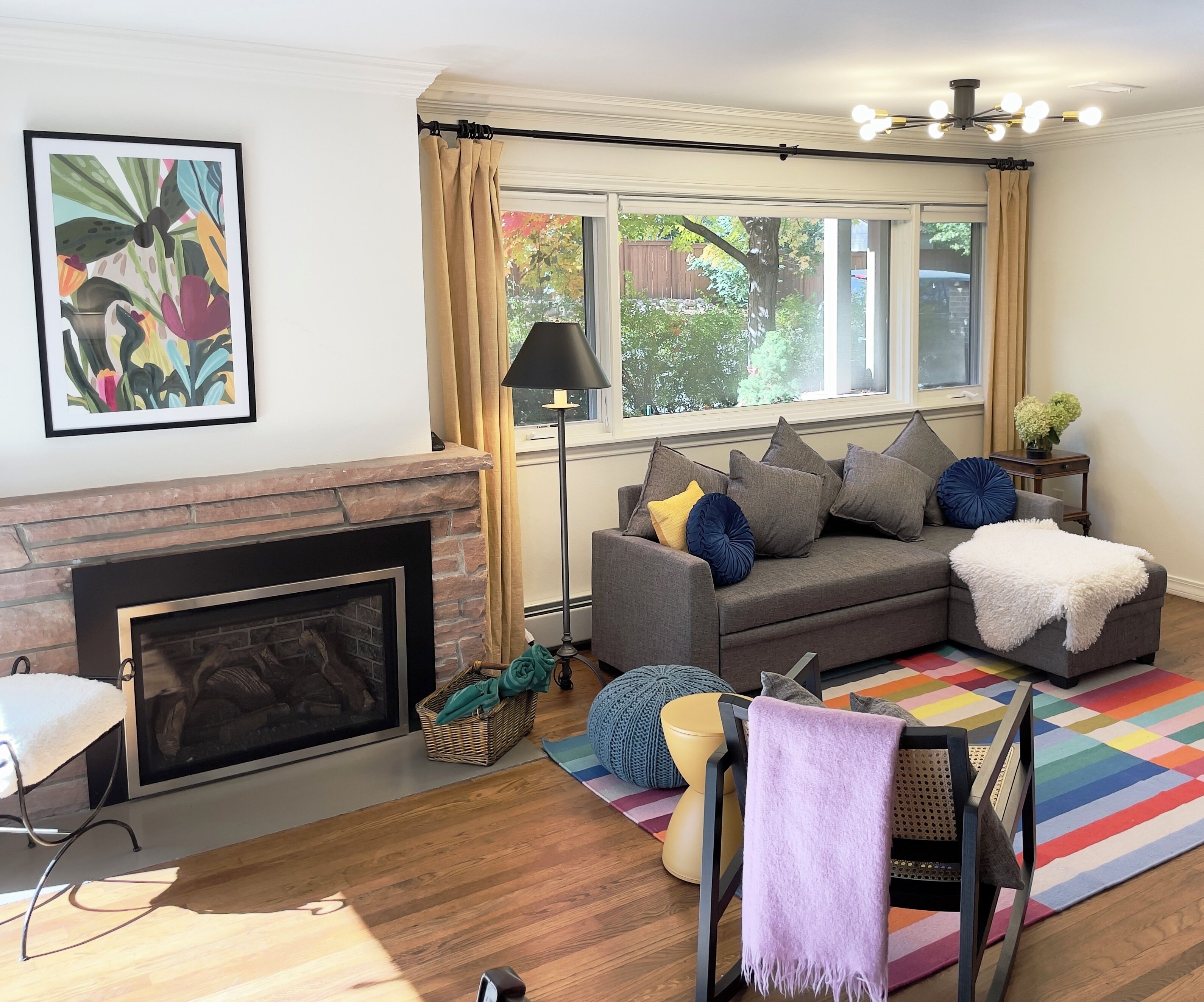 A living room with a fireplace, a gray sofa with cushions, a colorful rug, and a window with curtains.
