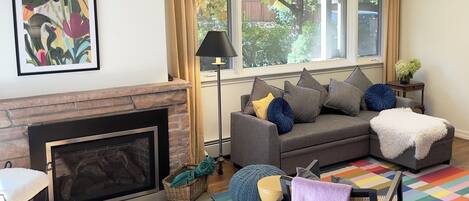 A living room with a fireplace, a gray sofa with cushions, a colorful rug, and a window with curtains.