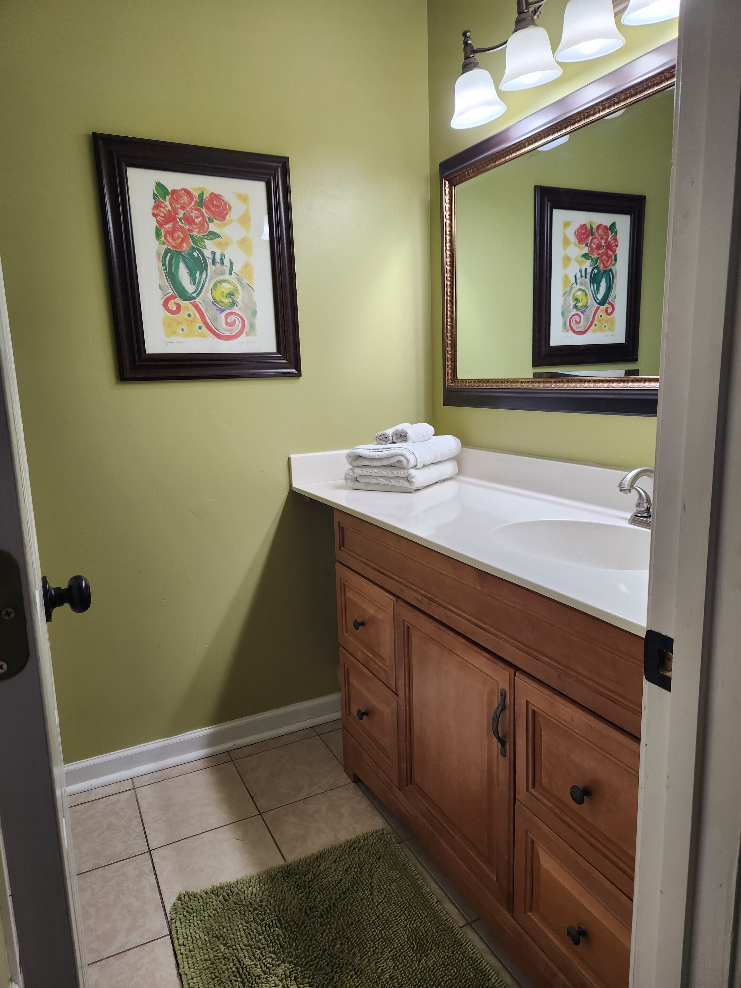 A bathroom with a white sink, wooden vanity, green walls, and a framed floral artwork above the mirror.