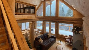 A spacious cabin interior with a wooden staircase, a cozy seating area with a sofa and chairs, and large windows offering views of the surrounding forest. - Douglas Fir Retreat | Mtn Views + Expansive Deck (Sunshine Valley)