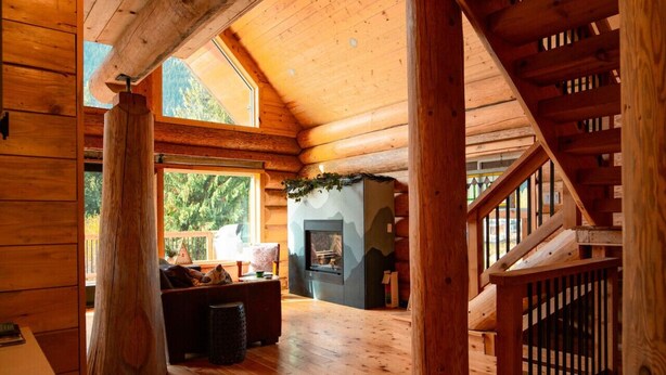 A log cabin interior with a fireplace, wooden staircase, and a cozy seating area. - Douglas Fir Retreat | Mtn Views + Expansive Deck (Sunshine Valley)