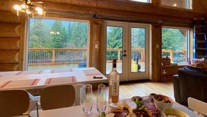 A wooden interior with a dining table set for a meal, featuring a charcuterie board, wine bottle, and glasses. - Douglas Fir Retreat | Mtn Views + Expansive Deck (Sunshine Valley)